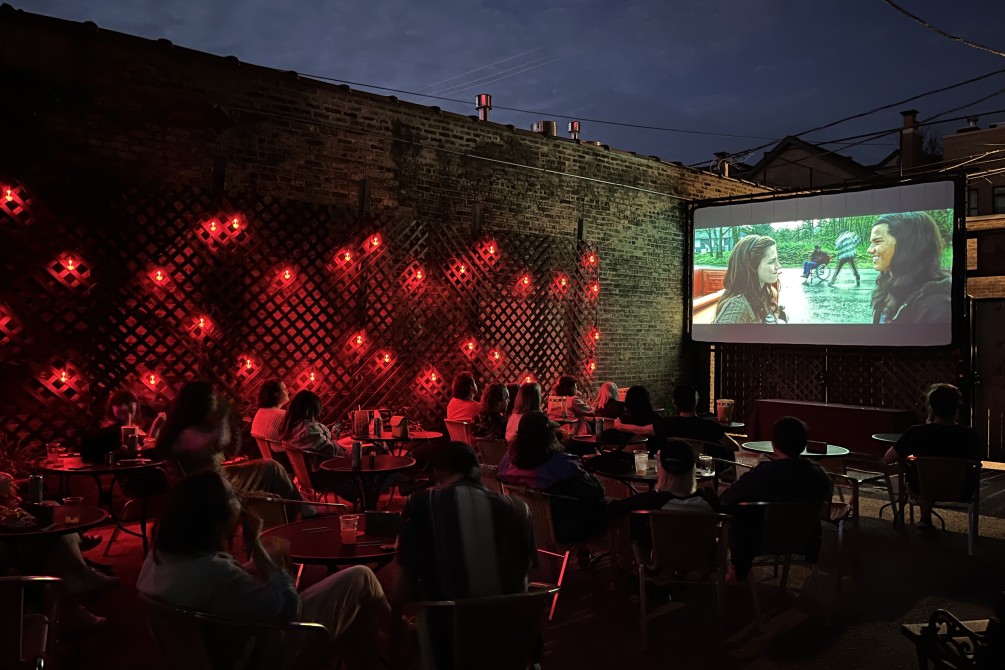 Outdoor seating at night with red lantern style lights strung along the brick wall and a movie screen in the background.