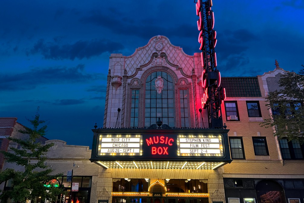 Dramatic 2022 photo of Music Box Theatre's exterior with a cloudy evening sky, bright marquee lights and a red glow reflecting off the building's exterior from the large neon sign.